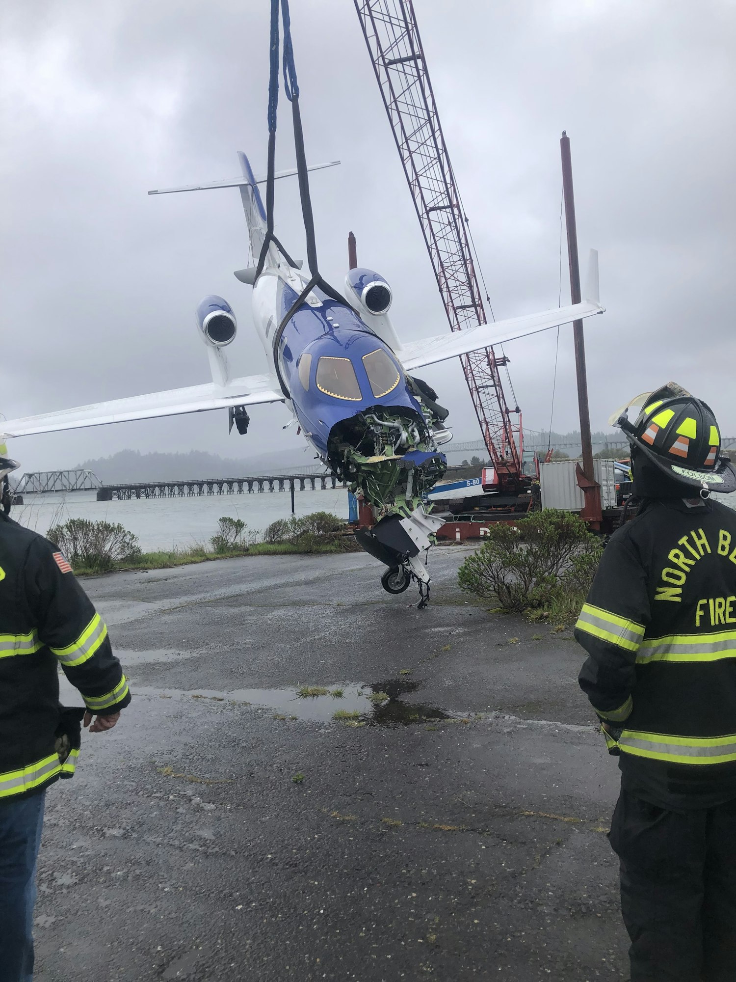 A small plane is being lifted by a crane, with firefighters standing nearby.