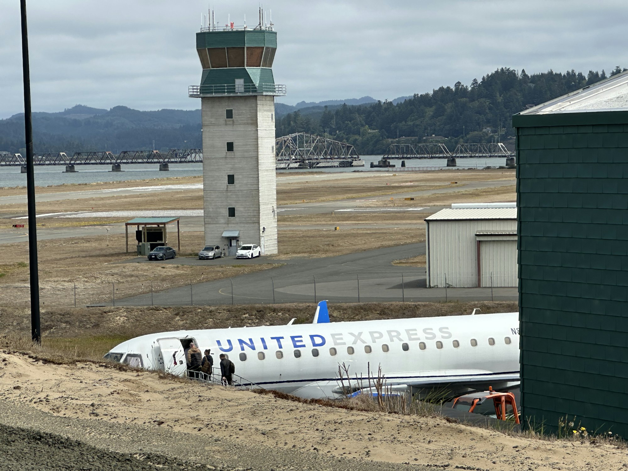 A United Express plane by an airport control tower, bridge, and parked cars, with people boarding or disembarking.