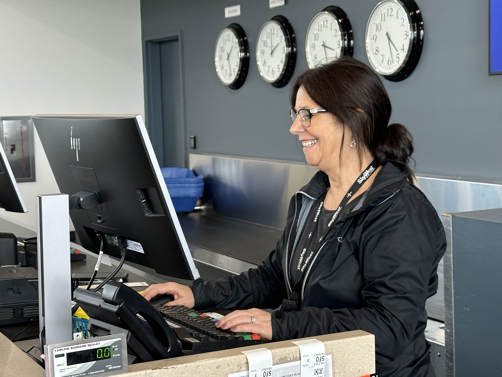 A smiling woman works at an airport check-in counter with clocks showing different time zones in the background.