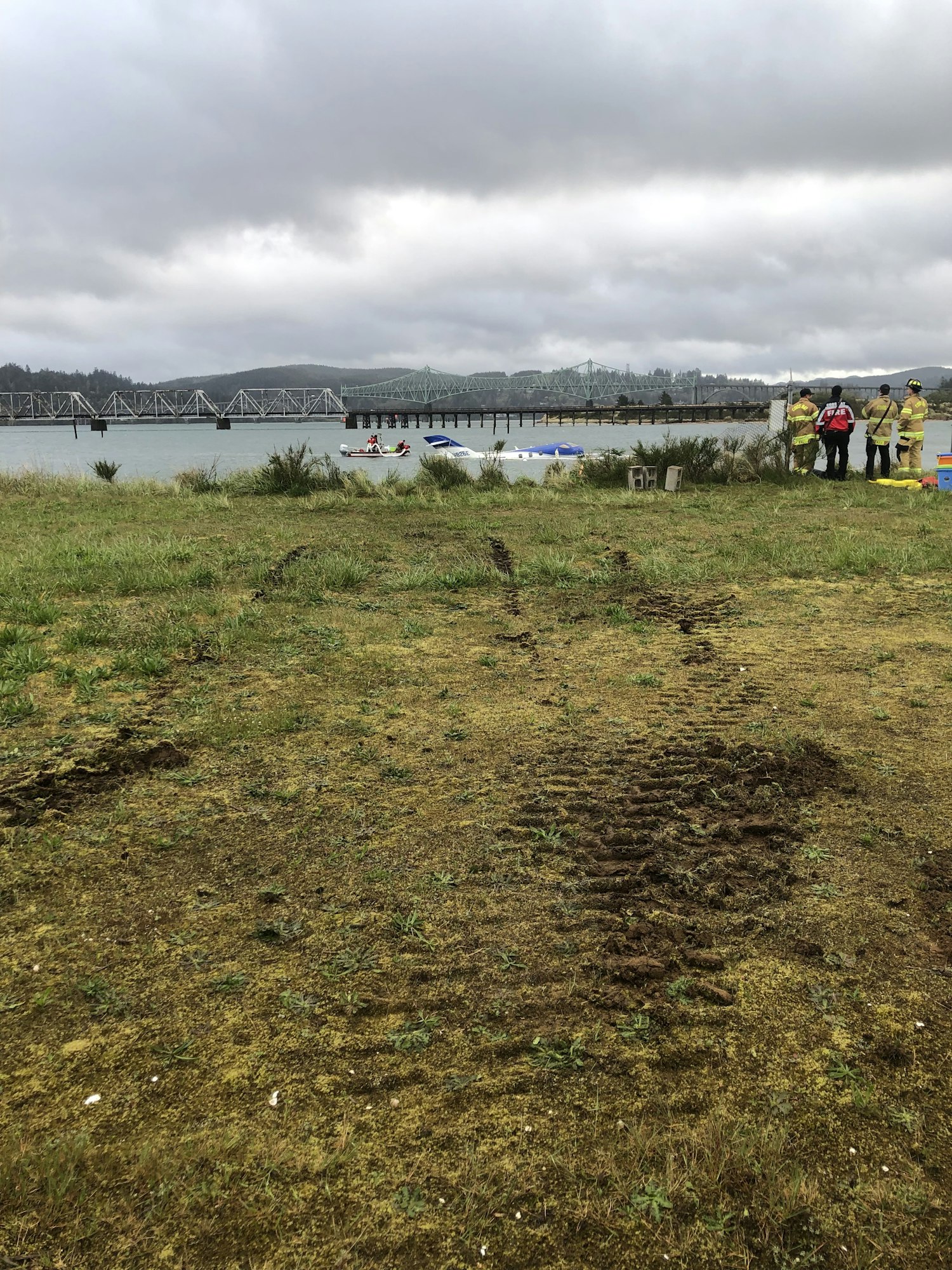 Grass, tire tracks, river, bridge, boats, and people in safety gear by the shore under a cloudy sky.