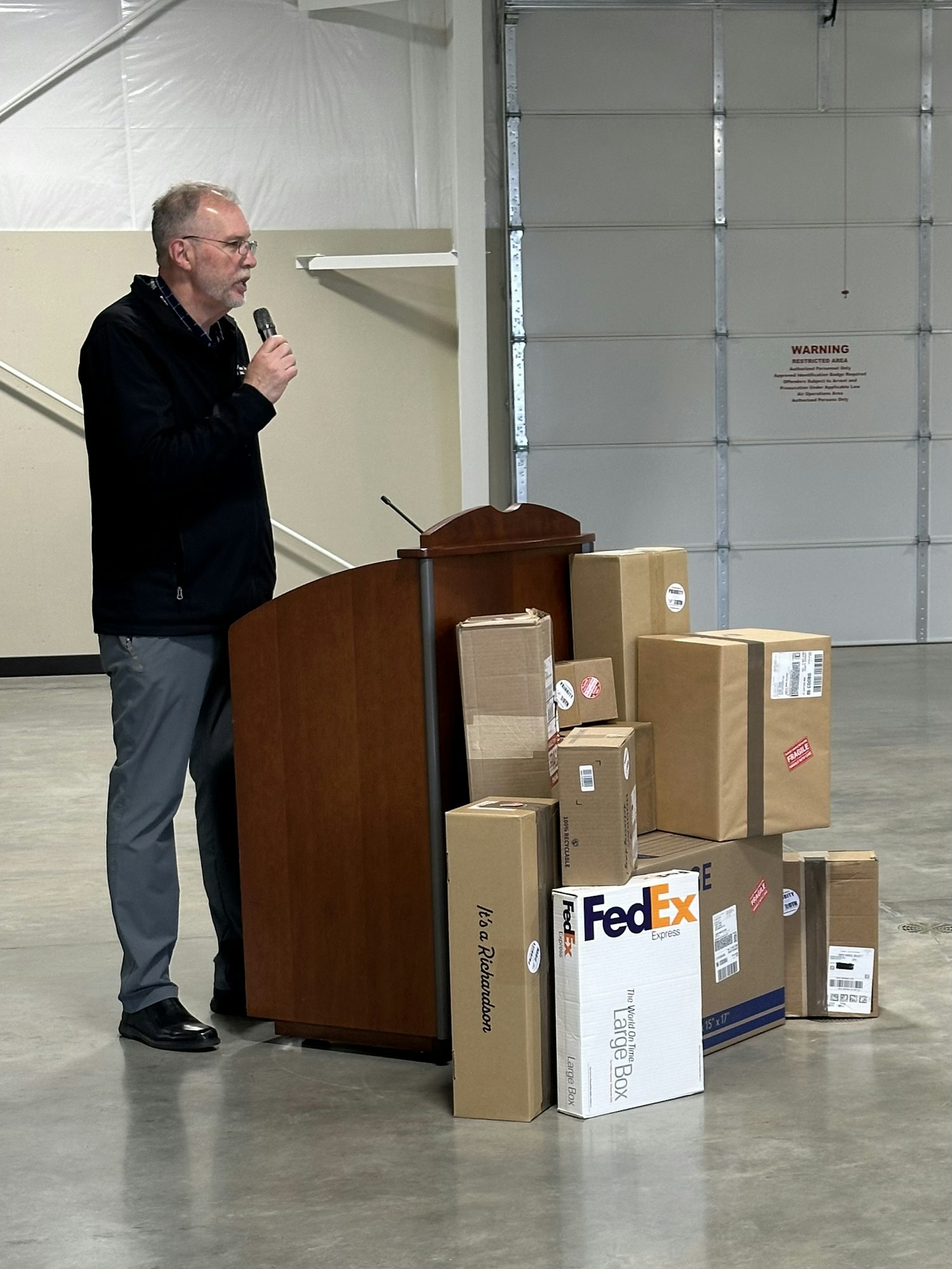 A speaker talks into a microphone beside a stack of packages, including FedEx boxes, in a large, empty room.