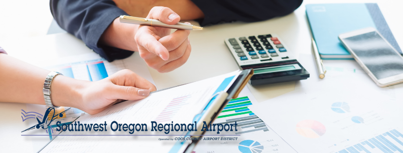 Person analyzing financial charts with pen, calculator & documents on desk. Logo: Southwest Oregon Regional Airport.