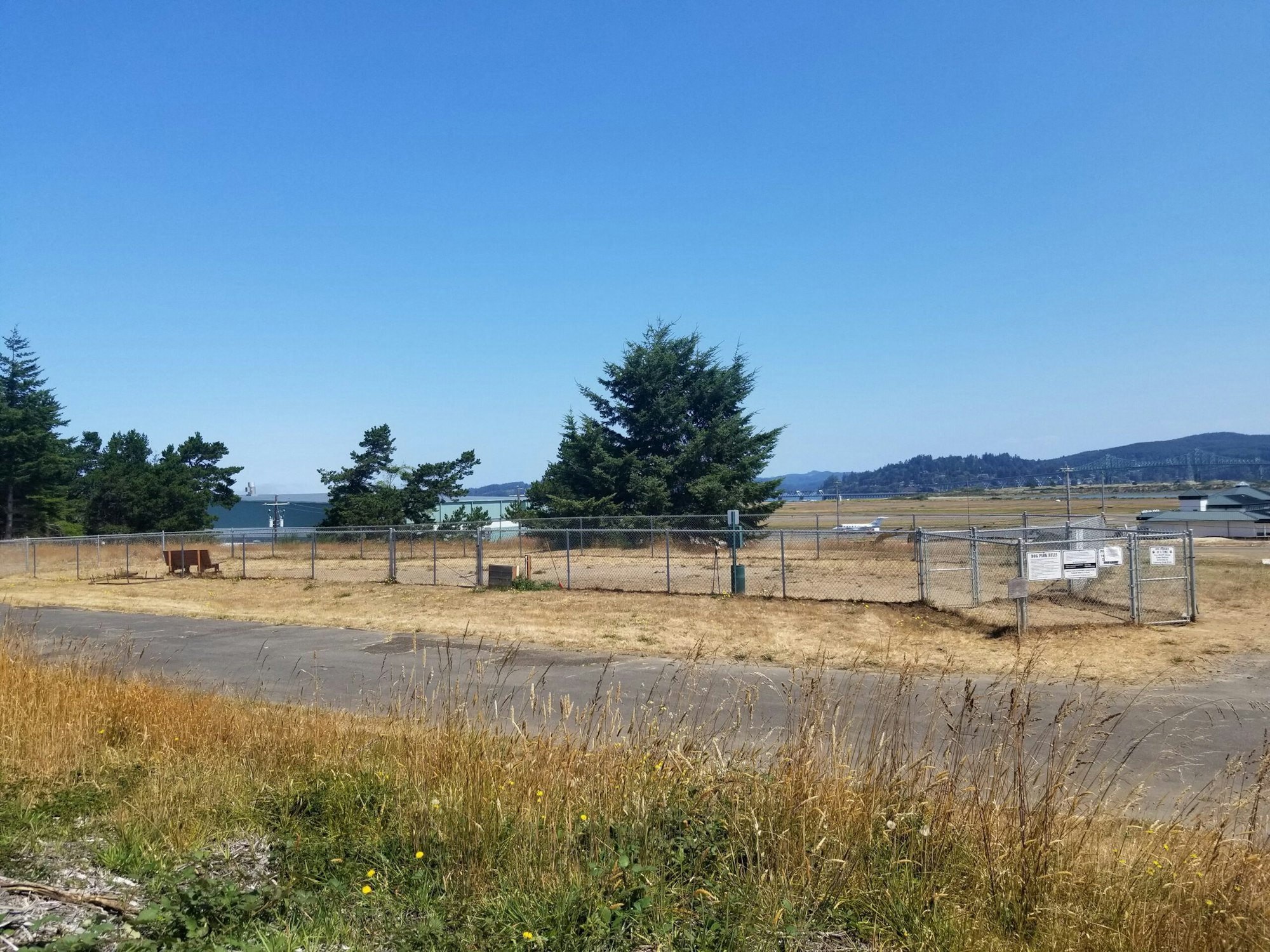 Rural airport with a chain-link fence, grassy field, trees, and a clear blue sky.