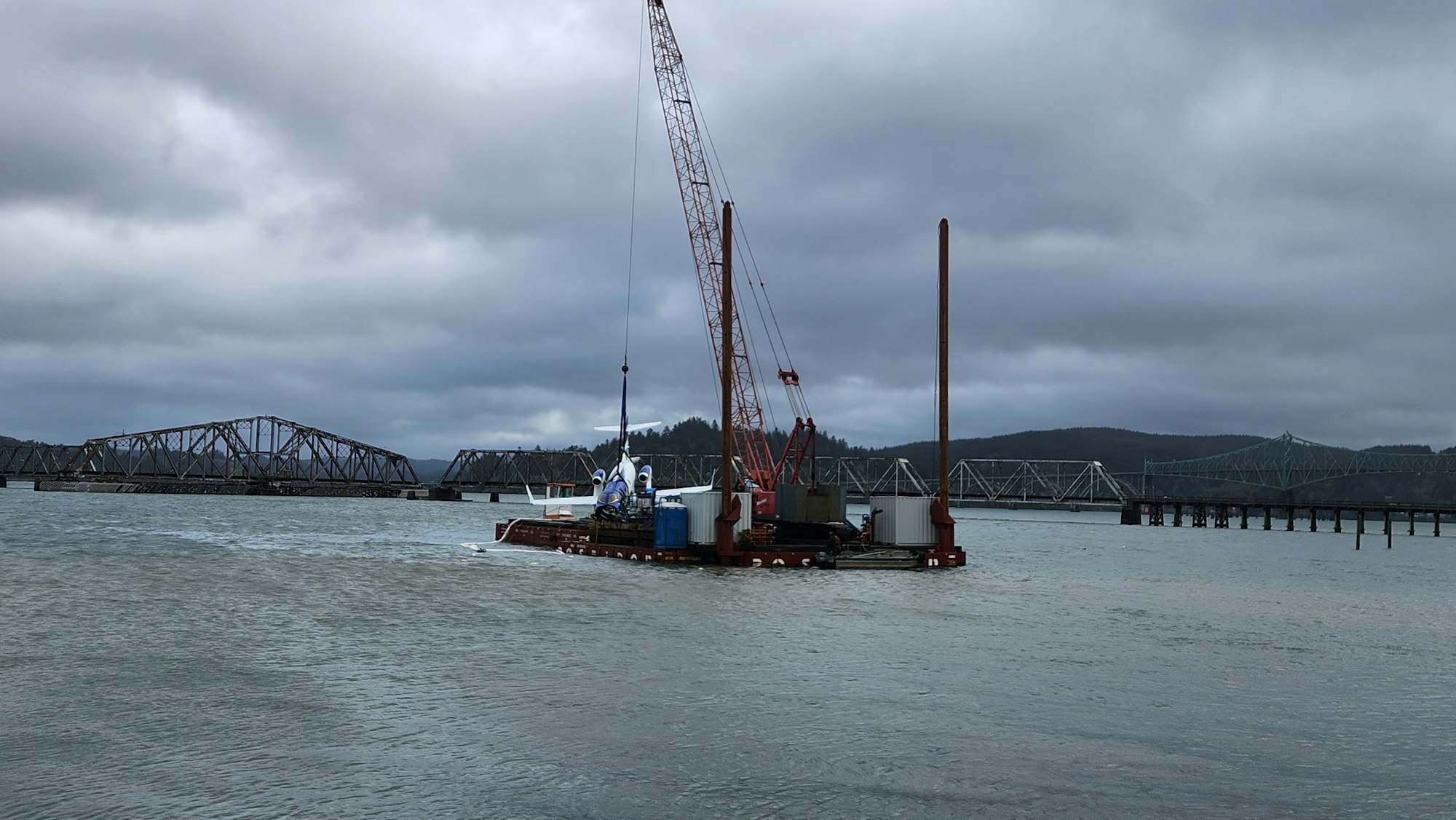 A crane barge on water with a bridge in the background under overcast skies.