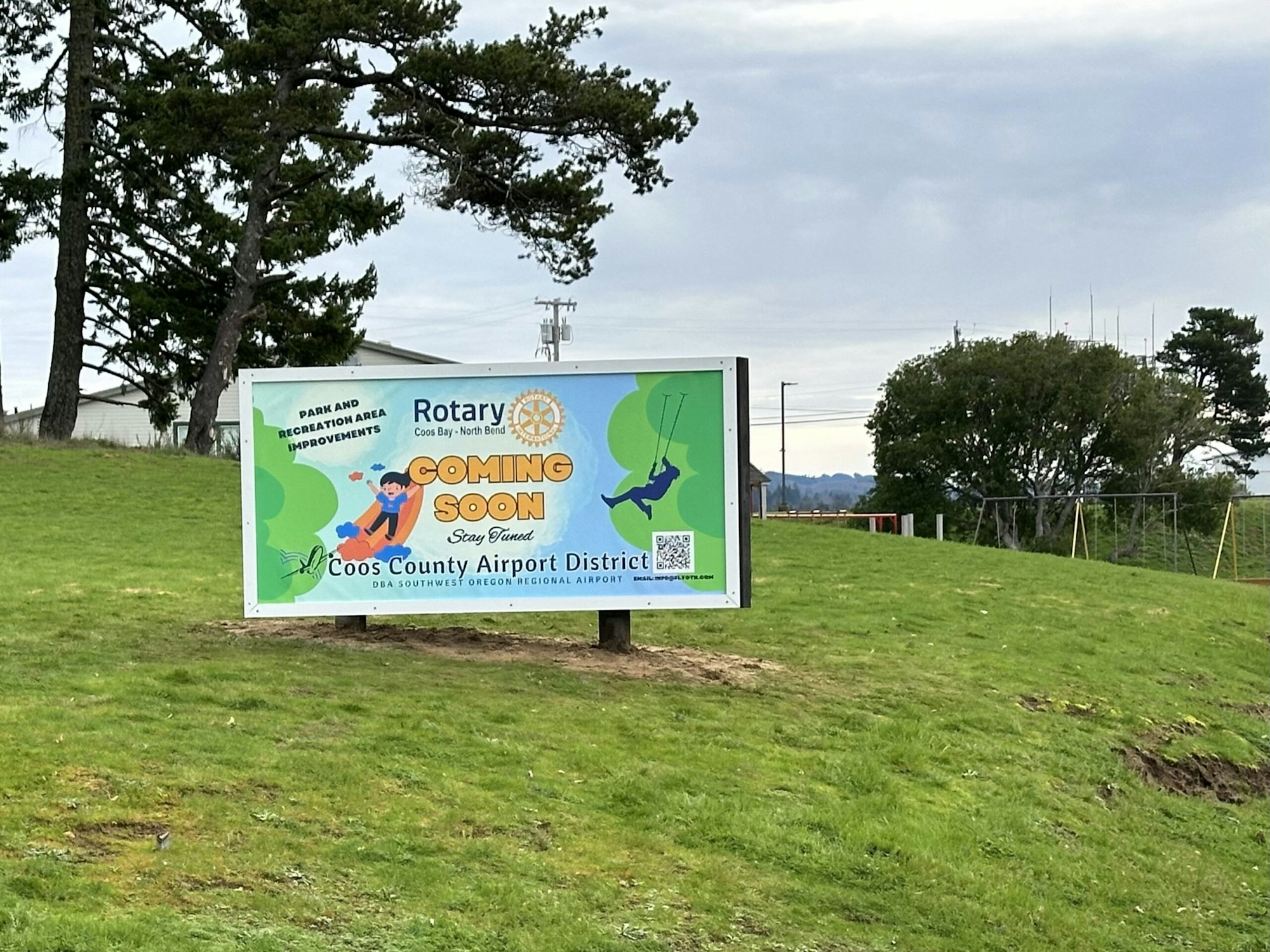 A billboard announcing "Coming Soon" park improvements by Rotary and Coos County Airport District, with greenery and cloudy skies in the background.
