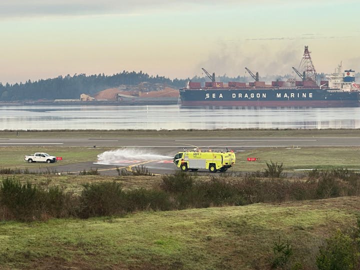 Airport fire truck spraying water, pickup truck on runway, cargo ship "SEA DRAGON MARINE", waterfront, hazy sky.