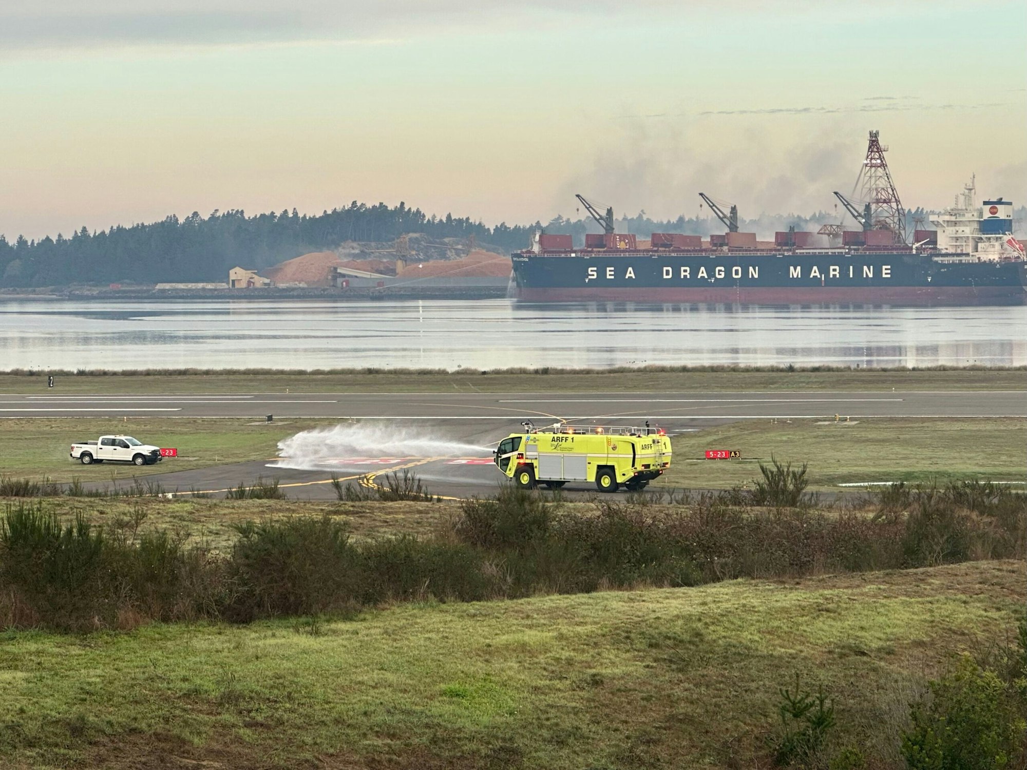 Airport fire truck spraying water, pickup truck on runway, cargo ship "SEA DRAGON MARINE", waterfront, hazy sky.