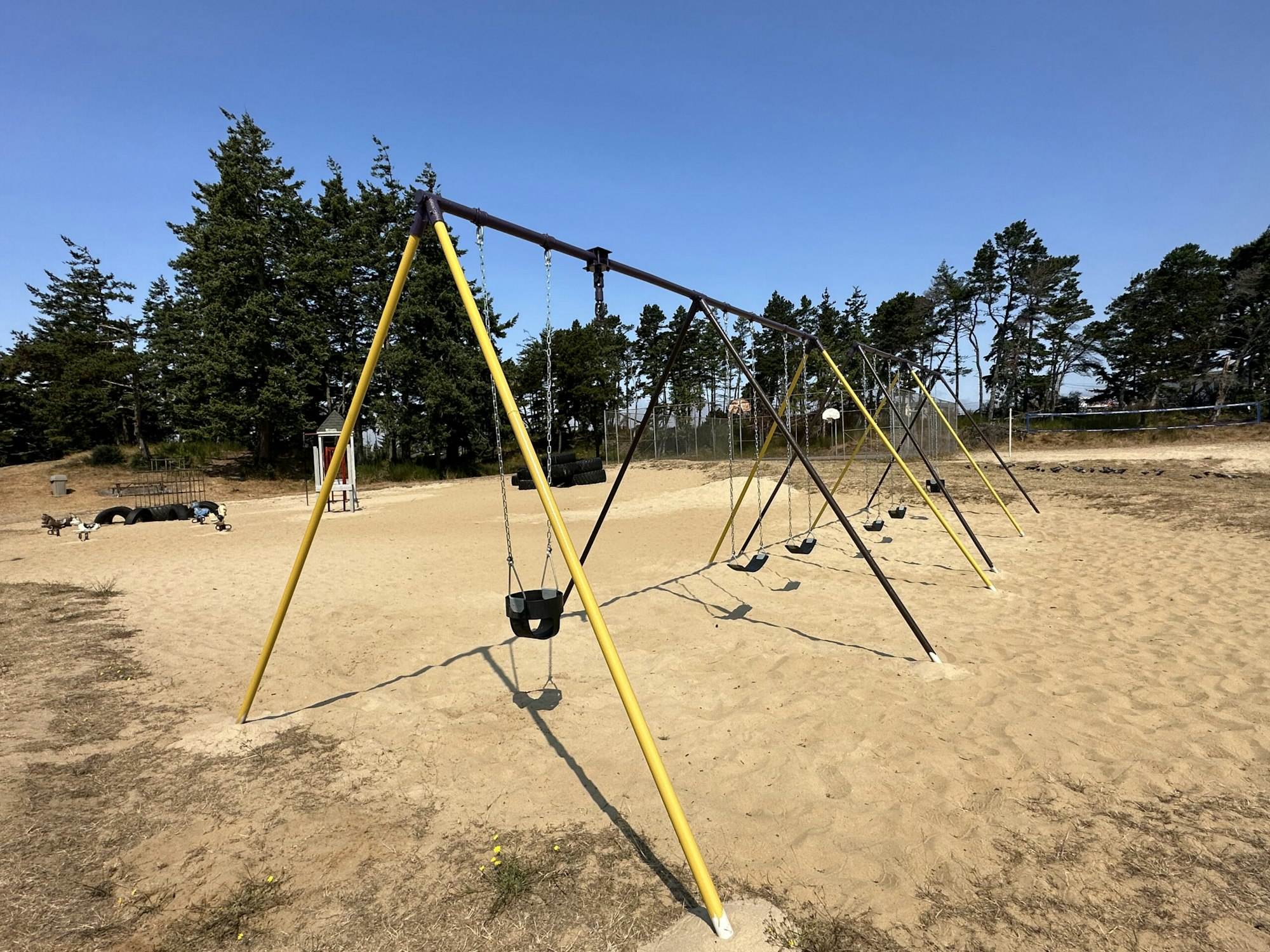 A playground with swings, sand, trees in the background, and a clear blue sky.