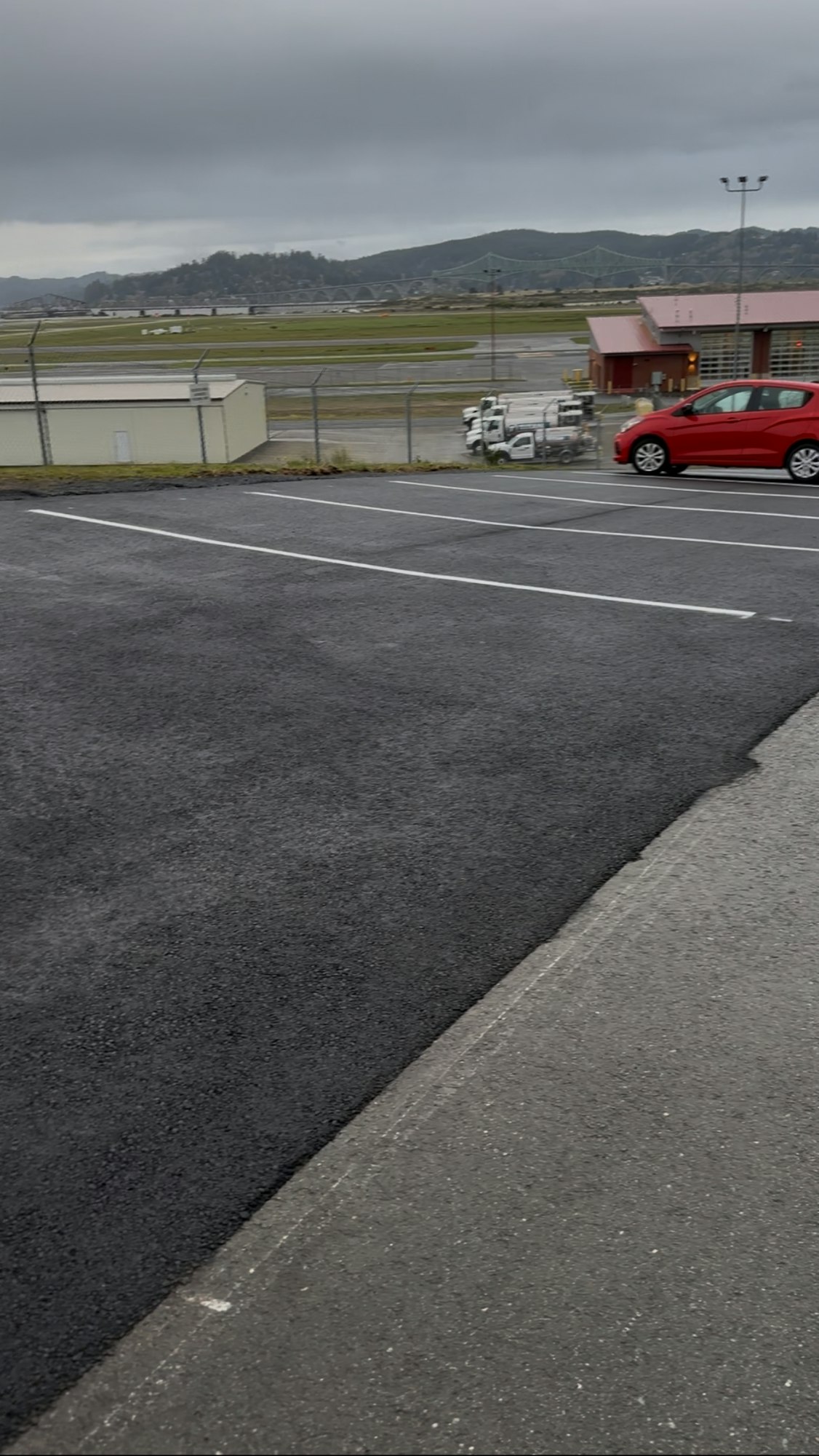 Parking lot with a red car, some white vehicles in the distance, a building, and a bridge against a cloudy sky.