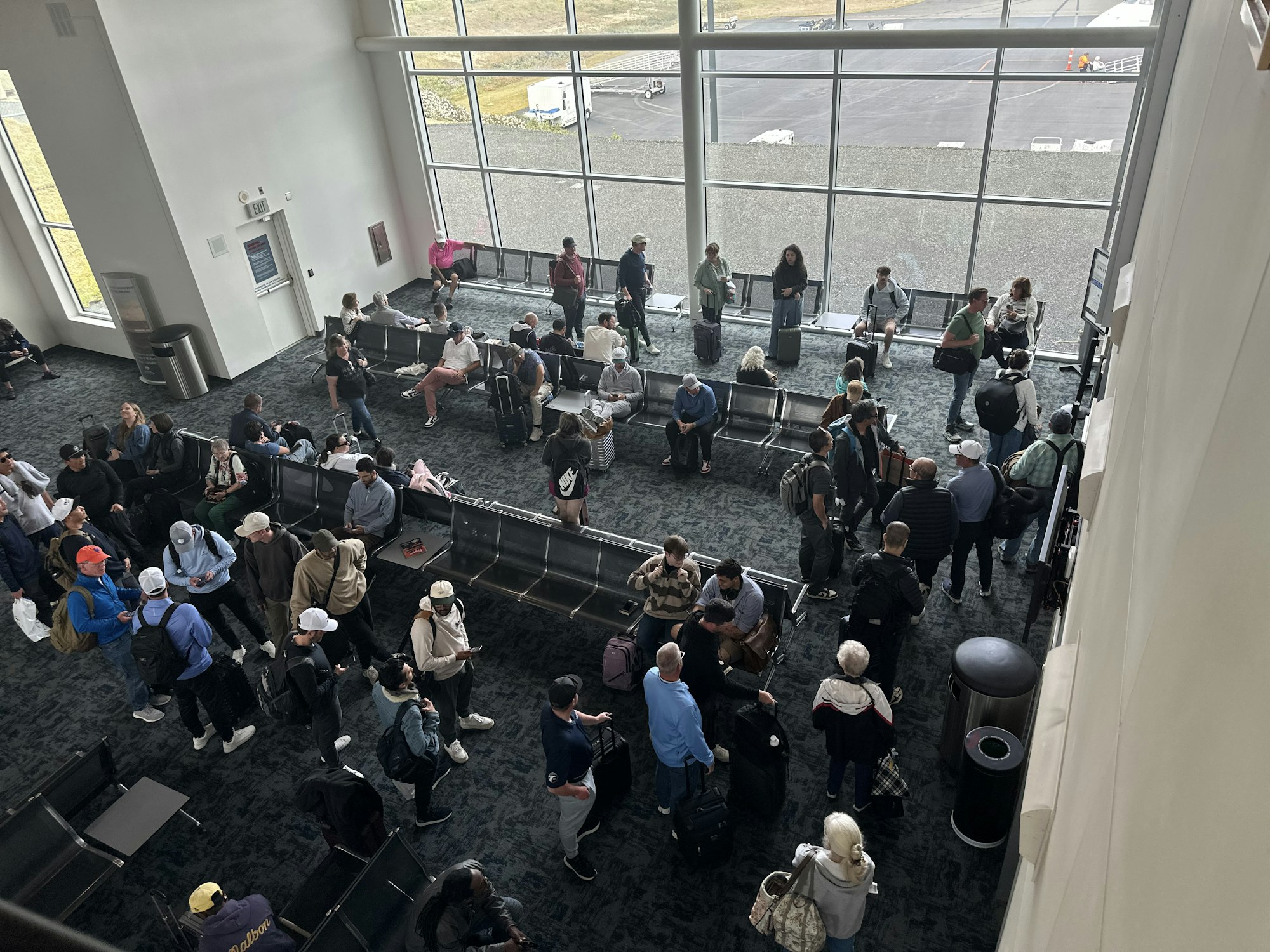 A busy airport waiting area with travelers seated and standing, some with luggage, near large windows.