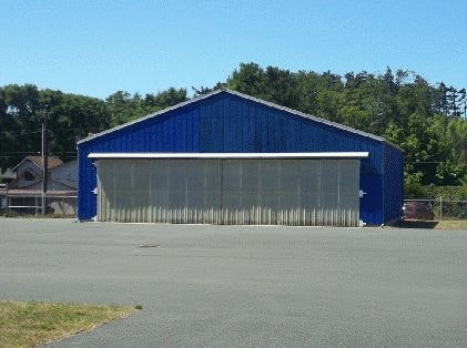 A blue industrial building with a large grey door, surrounded by a parking lot and trees in the background.