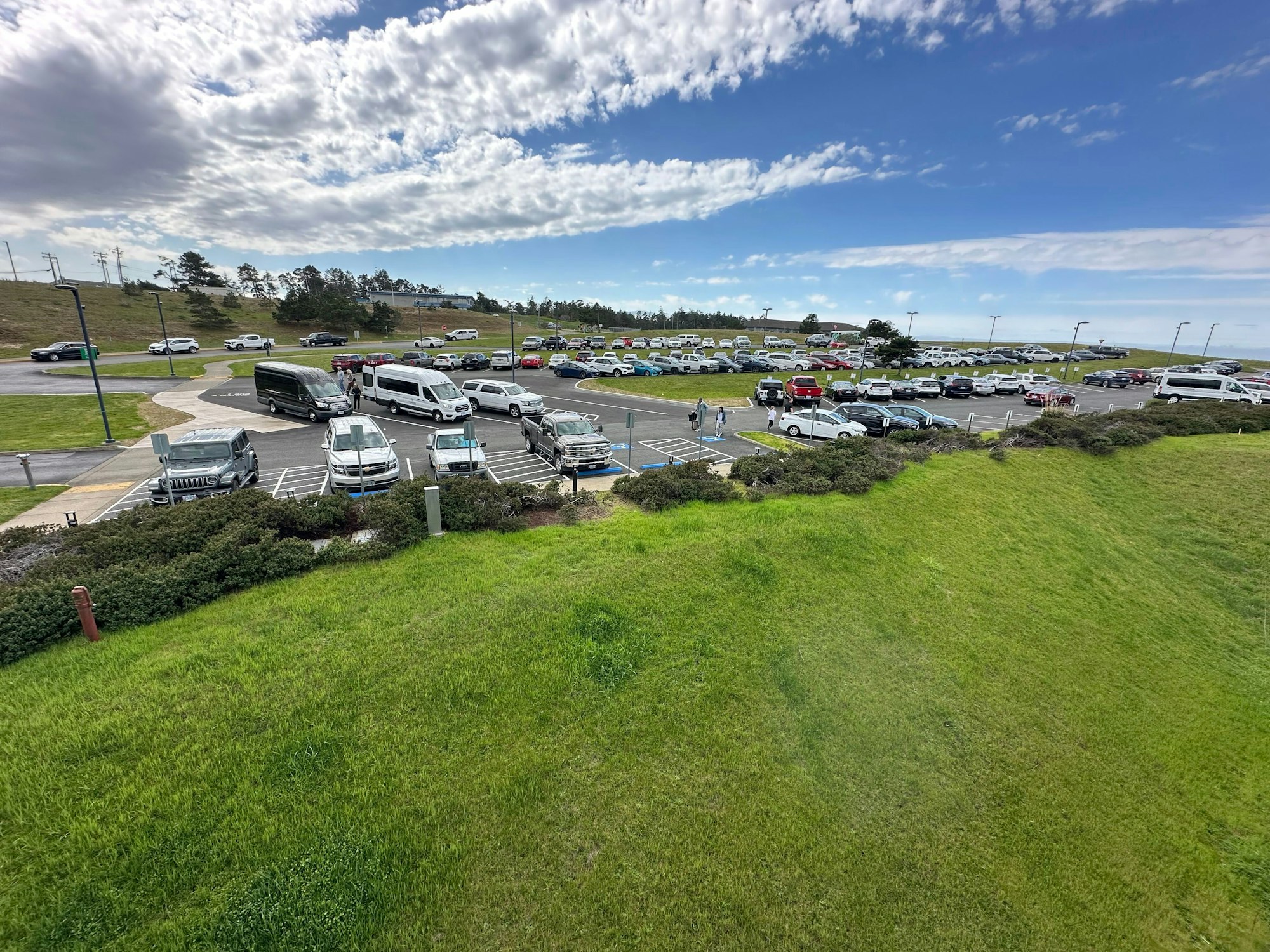 Parking lot with various cars, vans, and a bus set against a grassy hill and a partly cloudy sky.