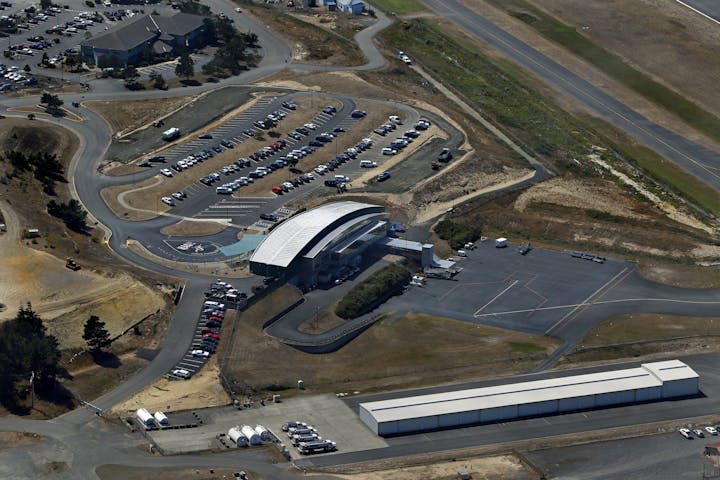 Aerial view of a small airport terminal with a curved roof, parking lot, and adjacent runway.