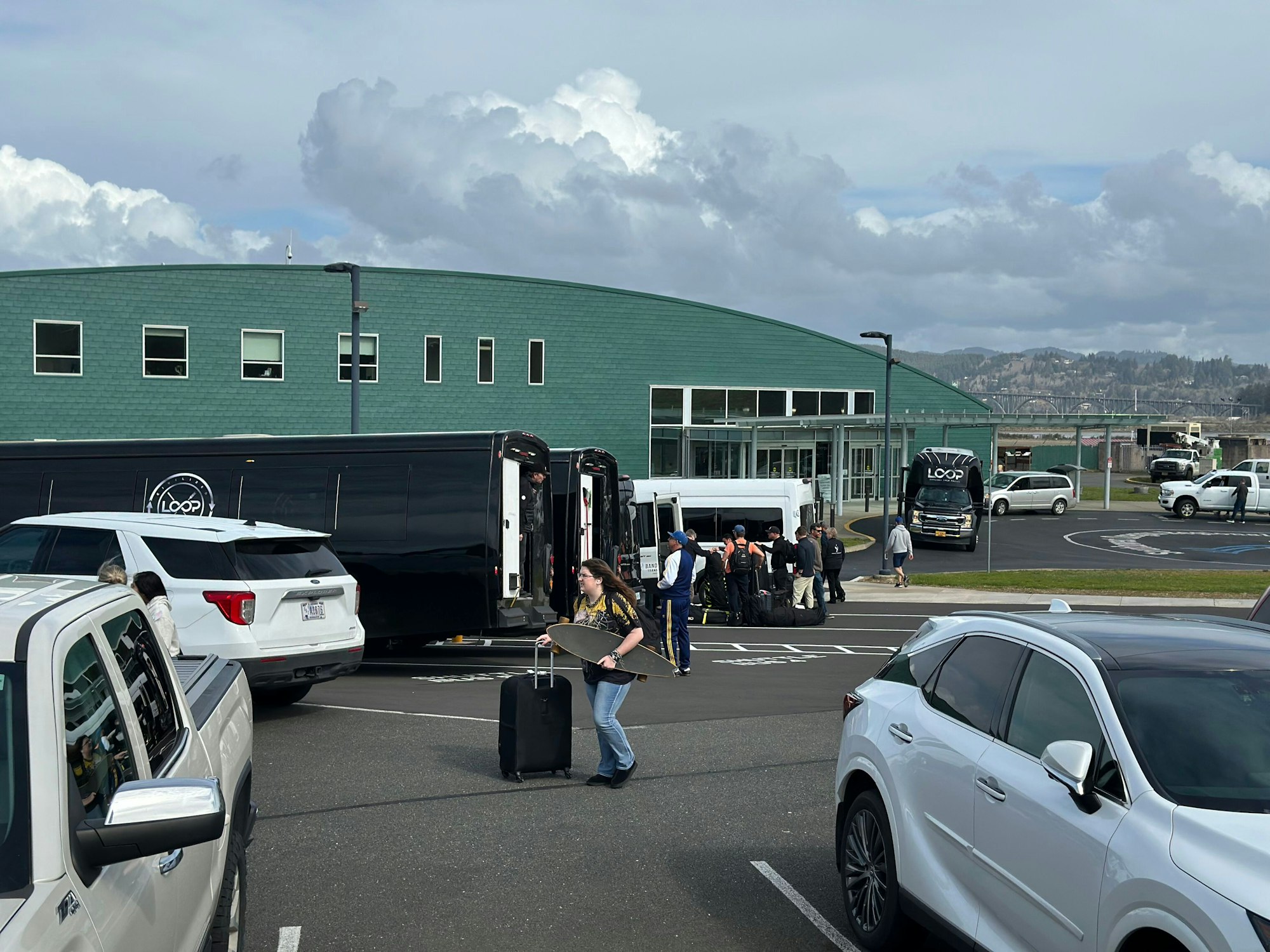 People with luggage near vans and cars in front of a green building under cloudy skies.