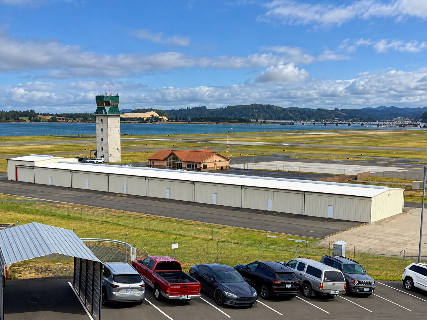The image shows the airport with a control tower, hangars, parked vehicles, and a scenic view of water and hills.