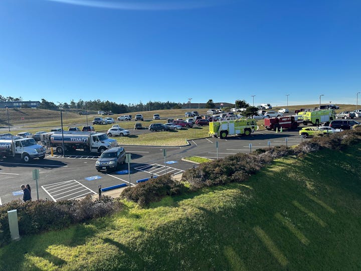 A parking lot with fire trucks, a tanker truck, and several other vehicles under a clear blue sky.
