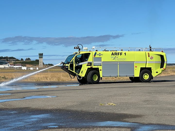 An airport firetruck spraying water on tarmac with a control tower in the background.