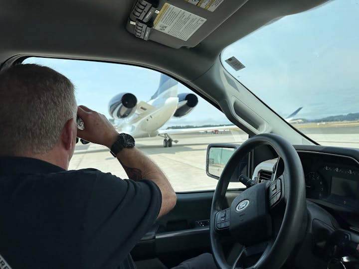 Person in a vehicle looking at an airplane on the tarmac through binoculars.
