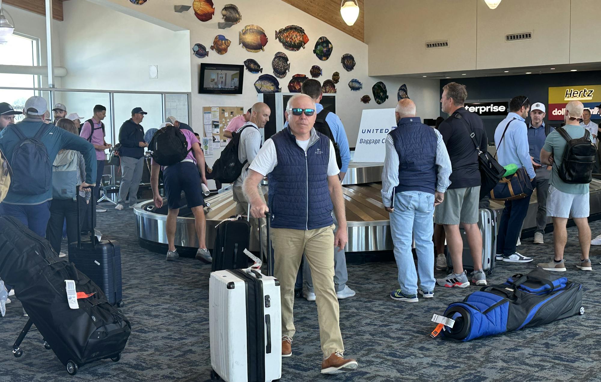 People at an airport baggage claim, some with luggage and golf bags, surrounded by wall decorations.