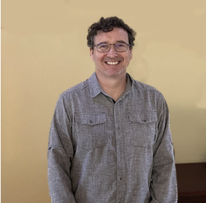 A smiling man wearing glasses and a gray shirt against a yellow wall.