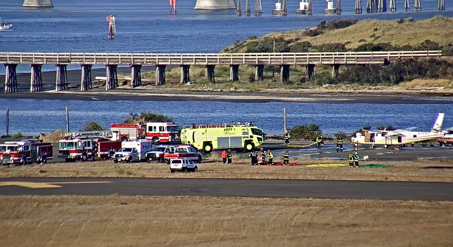 Emergency services at a runway with a small aircraft, near water and a bridge.