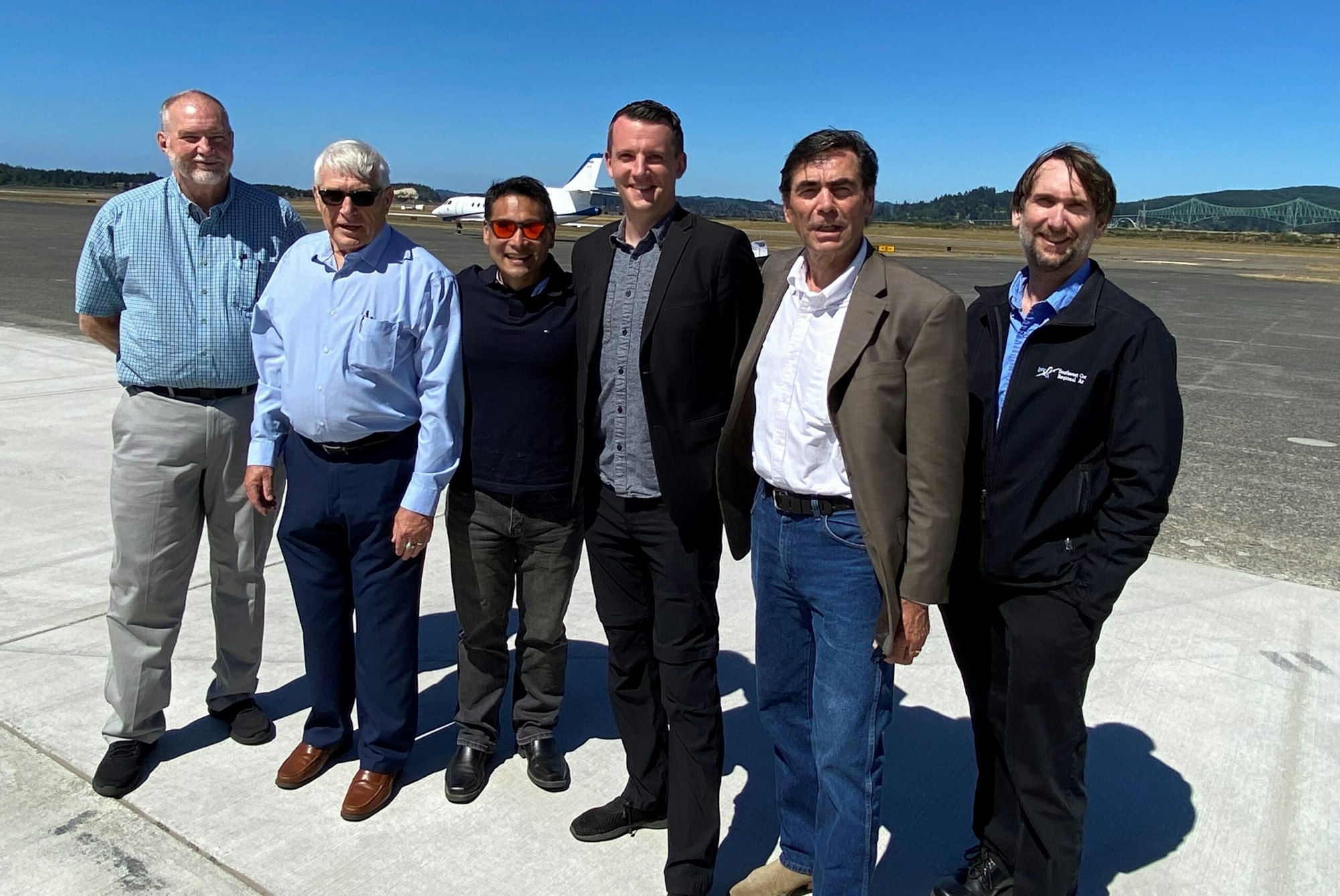 Six men standing on an airport tarmac with a jet and bridge in the background.