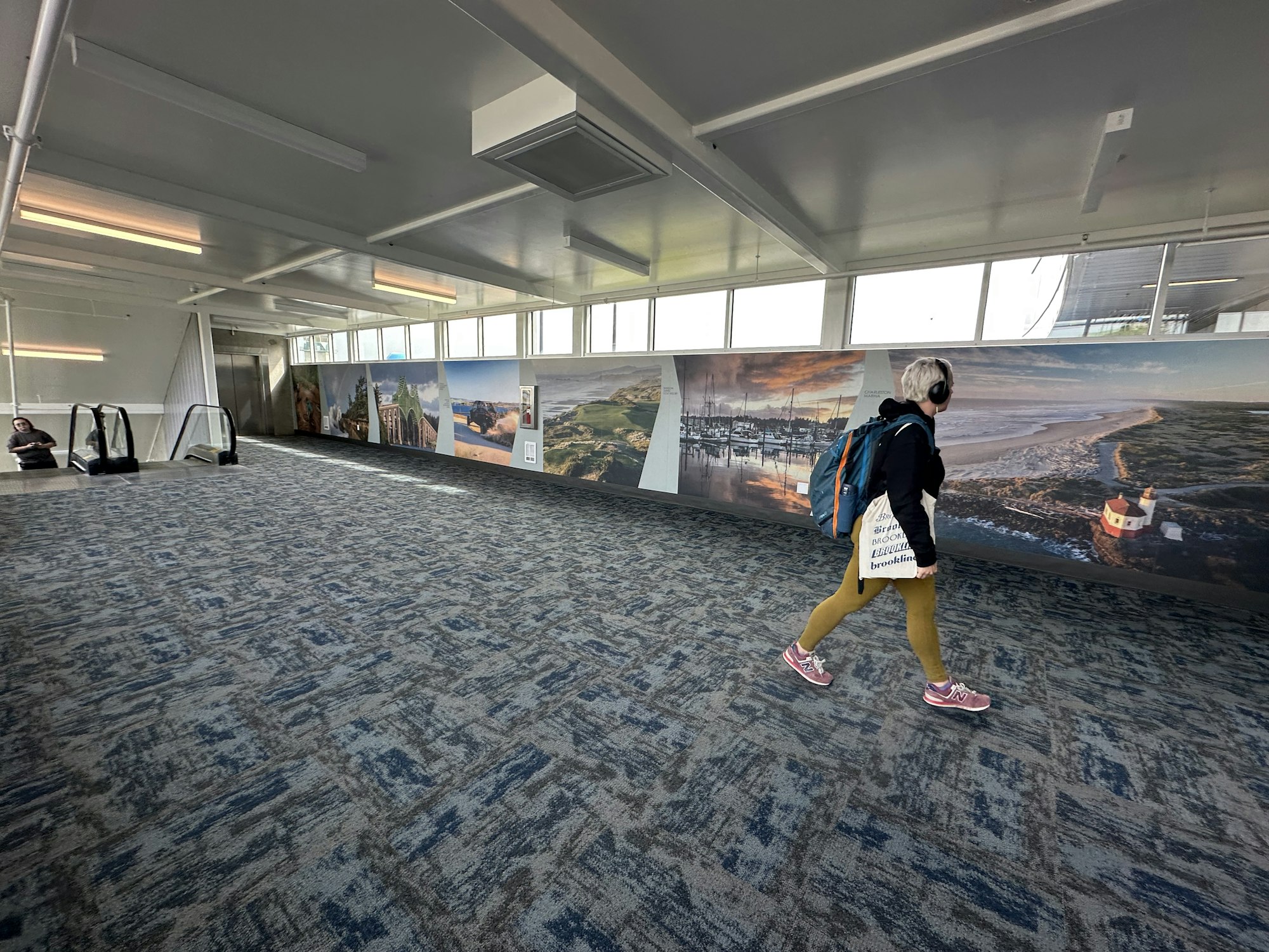 A person walks past a mural in a spacious area, with escalators nearby and windows letting in natural light.