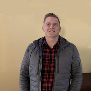 A smiling person wearing a plaid shirt and a jacket, standing in front of a yellow wall.