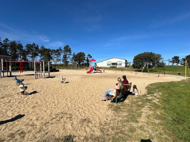 A sunny playground with swings, slide, climbing frame, spring riders, and people sitting.