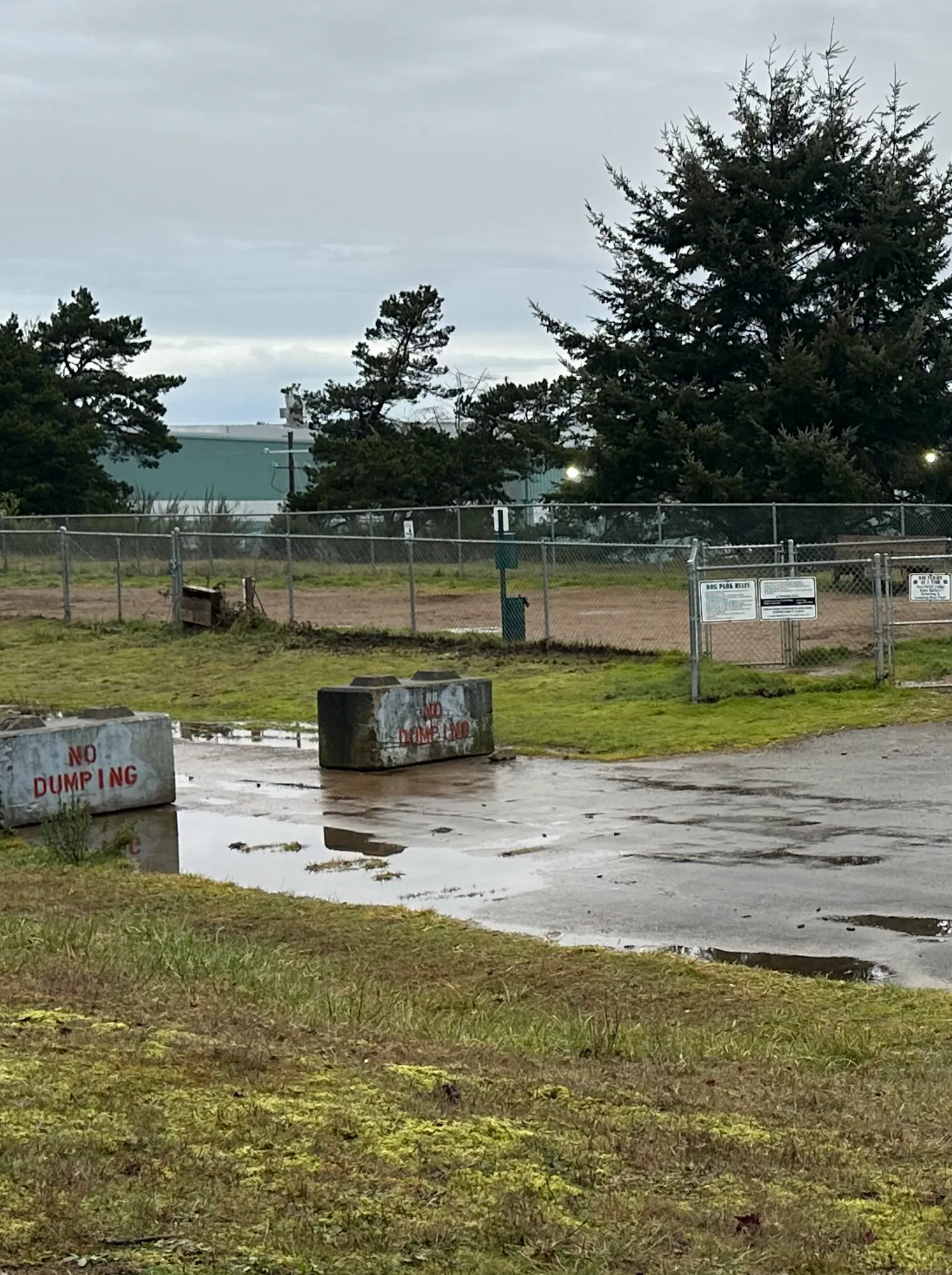 Dog Park Fenced area near runway with "No RV Parking" and "No Dumping" signs. Grass, trees, cloudy sky, and airplanes in the background.