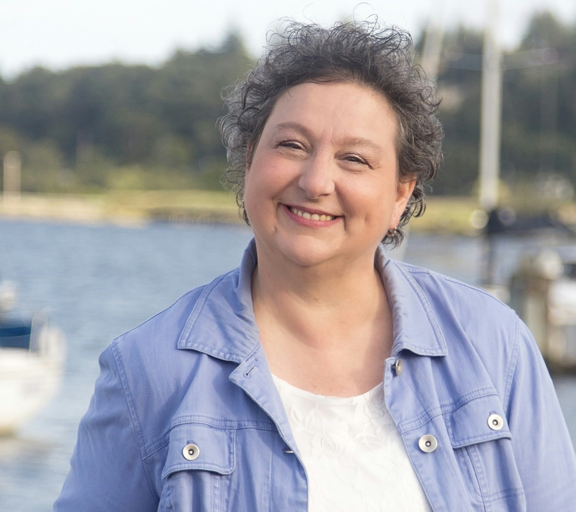 A smiling woman in a blue jacket and white top, with boats and water in the background.