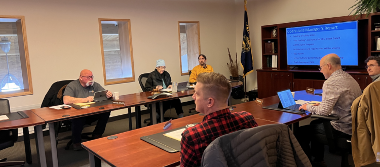 A business meeting with six people, laptops, documents, and a presentation in a conference room.