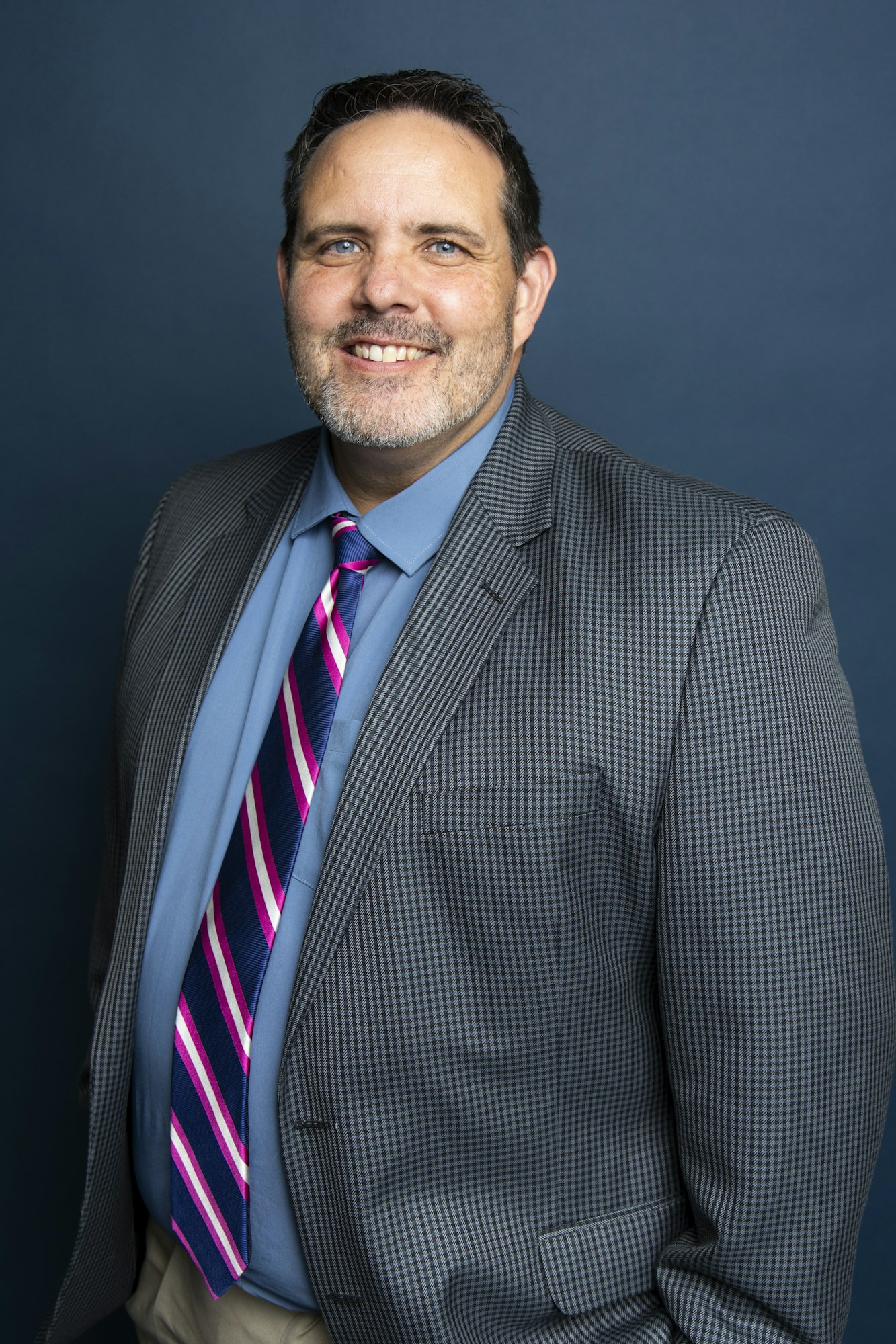 A person in a checkered suit and striped tie smiles against a blue background.