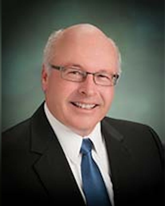 A smiling person with glasses wearing a dark suit, white shirt, and tie against a graduated backdrop.