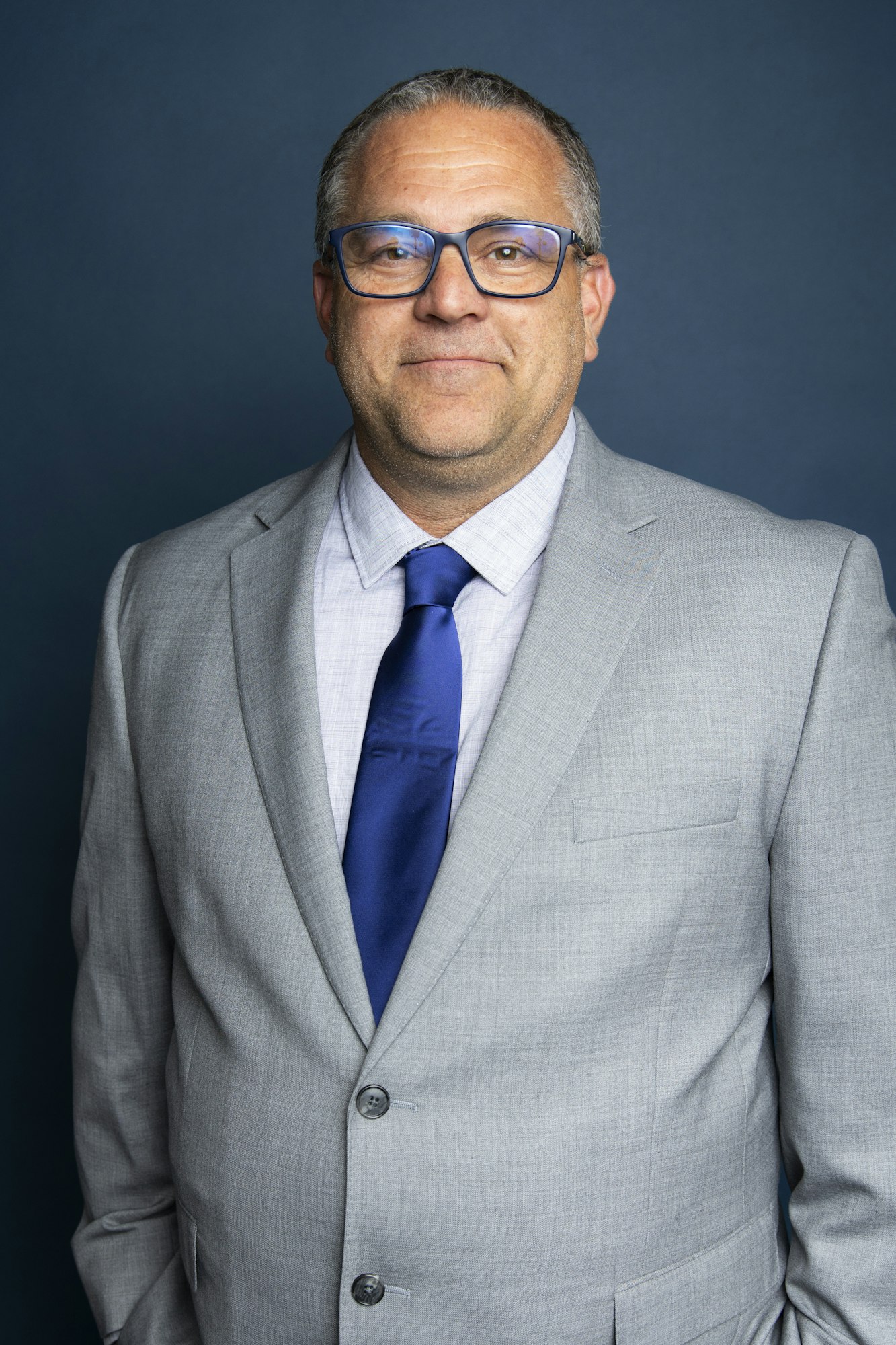 A man in a light gray suit with a blue tie and glasses stands against a dark background.