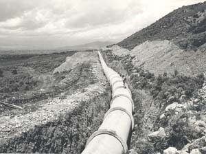 A long pipeline stretches through rough terrain under a cloudy sky.
