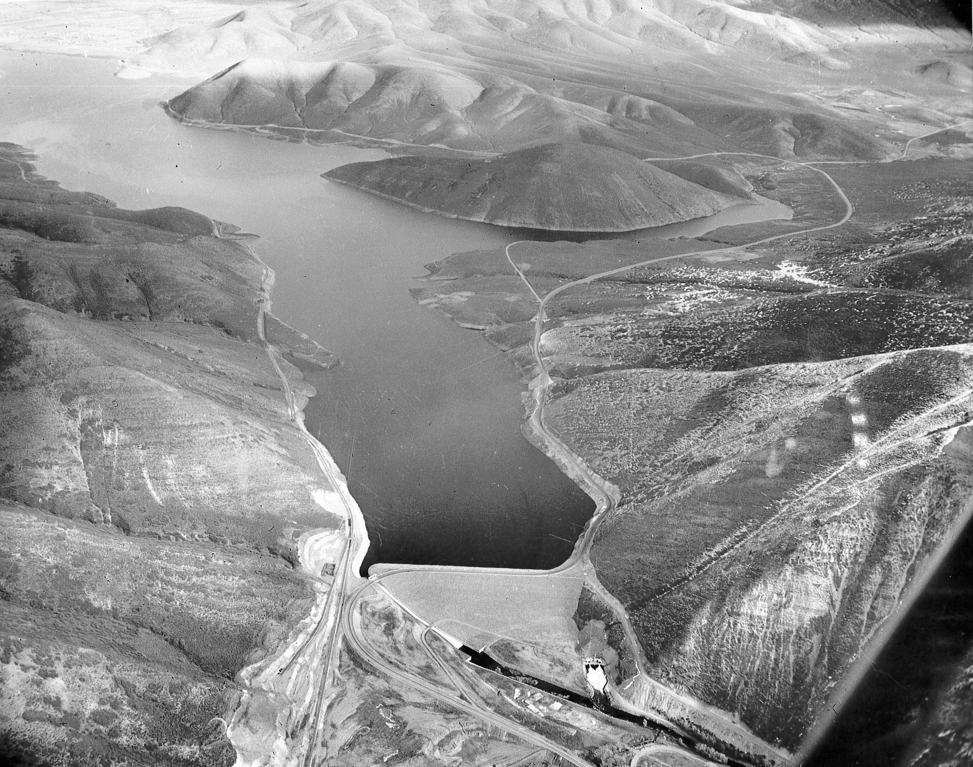The image shows a black-and-white aerial view of a river or reservoir, surrounded by hilly terrain and possibly infrastructure.
