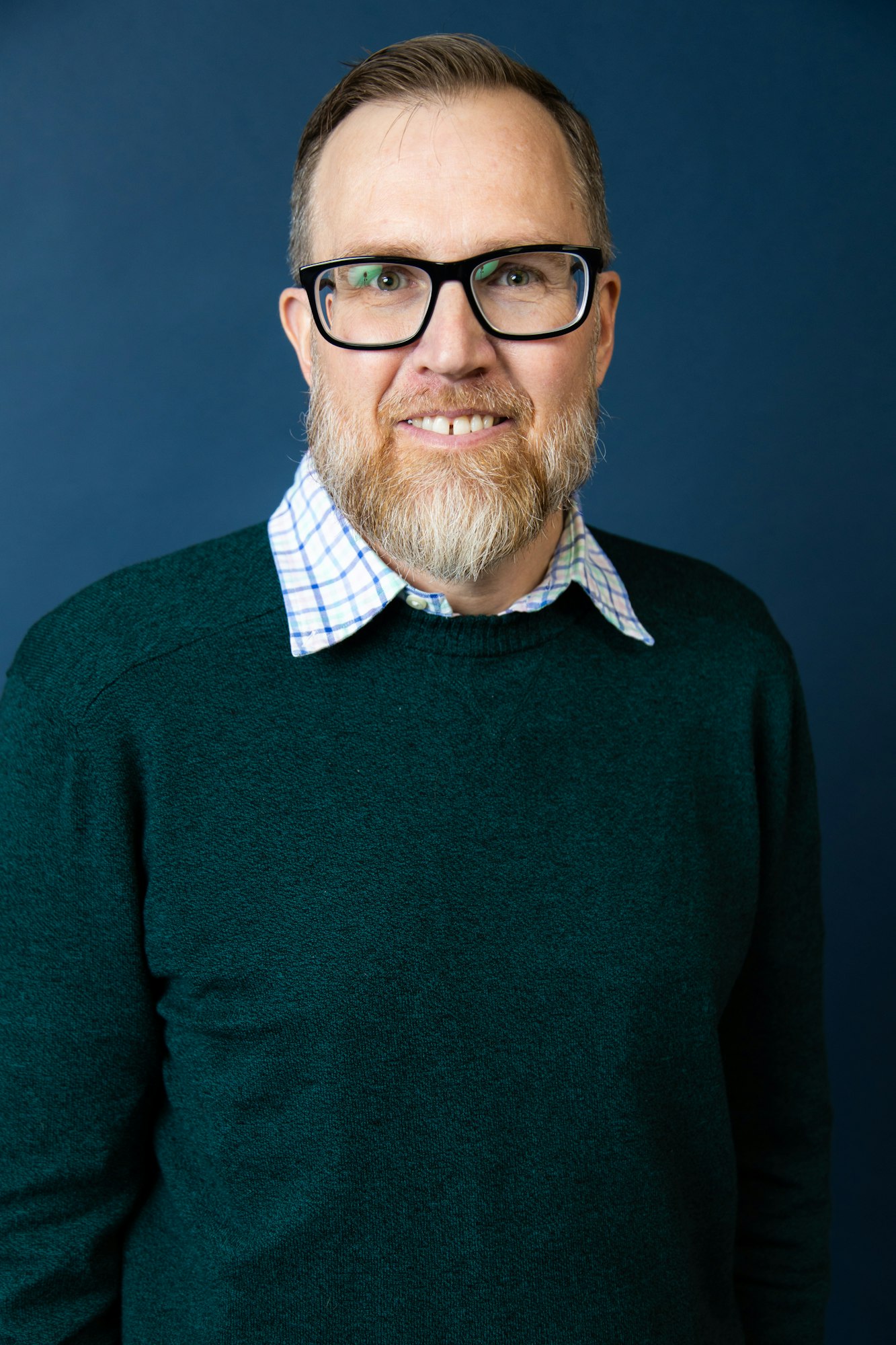 A smiling man with glasses, a beard, and a green sweater over a checked shirt against a blue background.