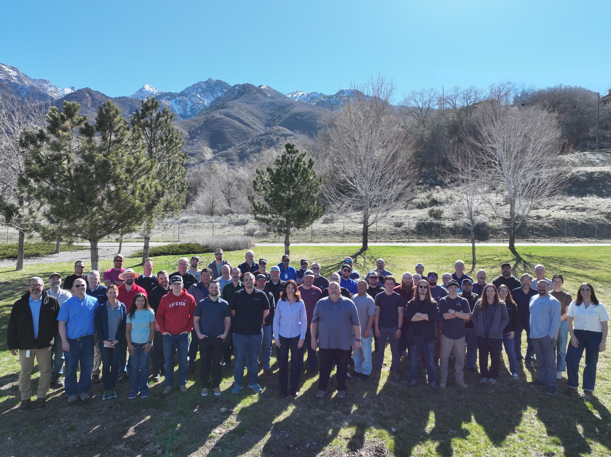 A group photo of about 50 people posing outdoors with mountains in the background, trees, and bright blue skies.
