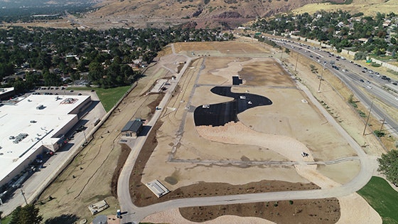 Aerial view of a landscape with paths, greenery, a large sandy area, and a unique building structure near a highway.