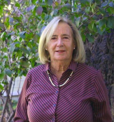 A person with blonde hair wearing a striped shirt and necklace stands outdoors in front of trees.