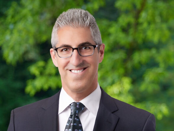 A person in a suit and tie, wearing glasses, smiling, with a blurred green foliage background.