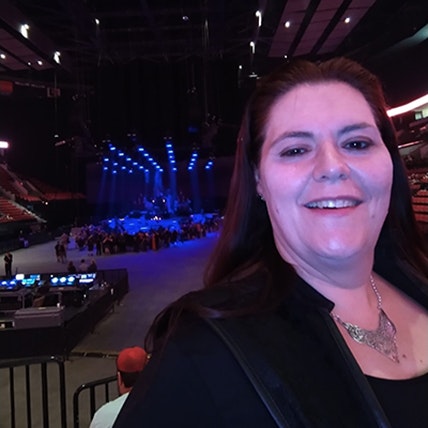 A person smiling at a concert venue with a stage and blue lights in the background.