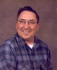A person is smiling, wearing glasses and a plaid shirt, against a plain background.
