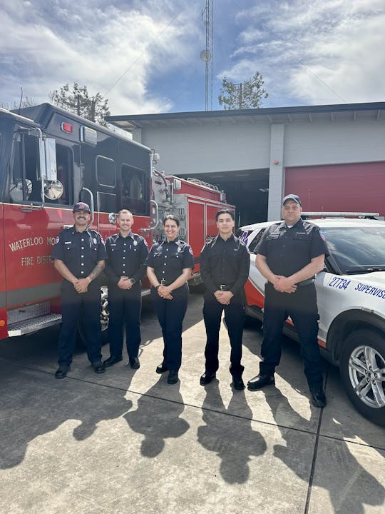 Five firefighters standing in front of fire trucks outside a station.
