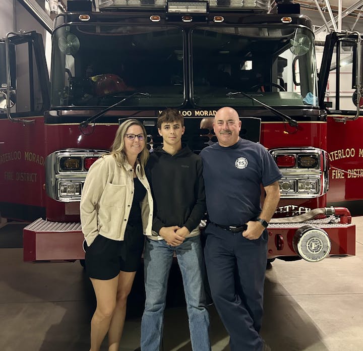 Two adults and a teenager standing in front of a red fire truck inside a fire station.