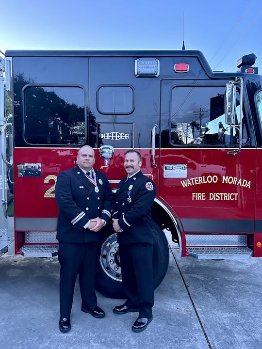 Two uniformed firefighters standing in front of a Waterloo Morada Fire District fire truck.