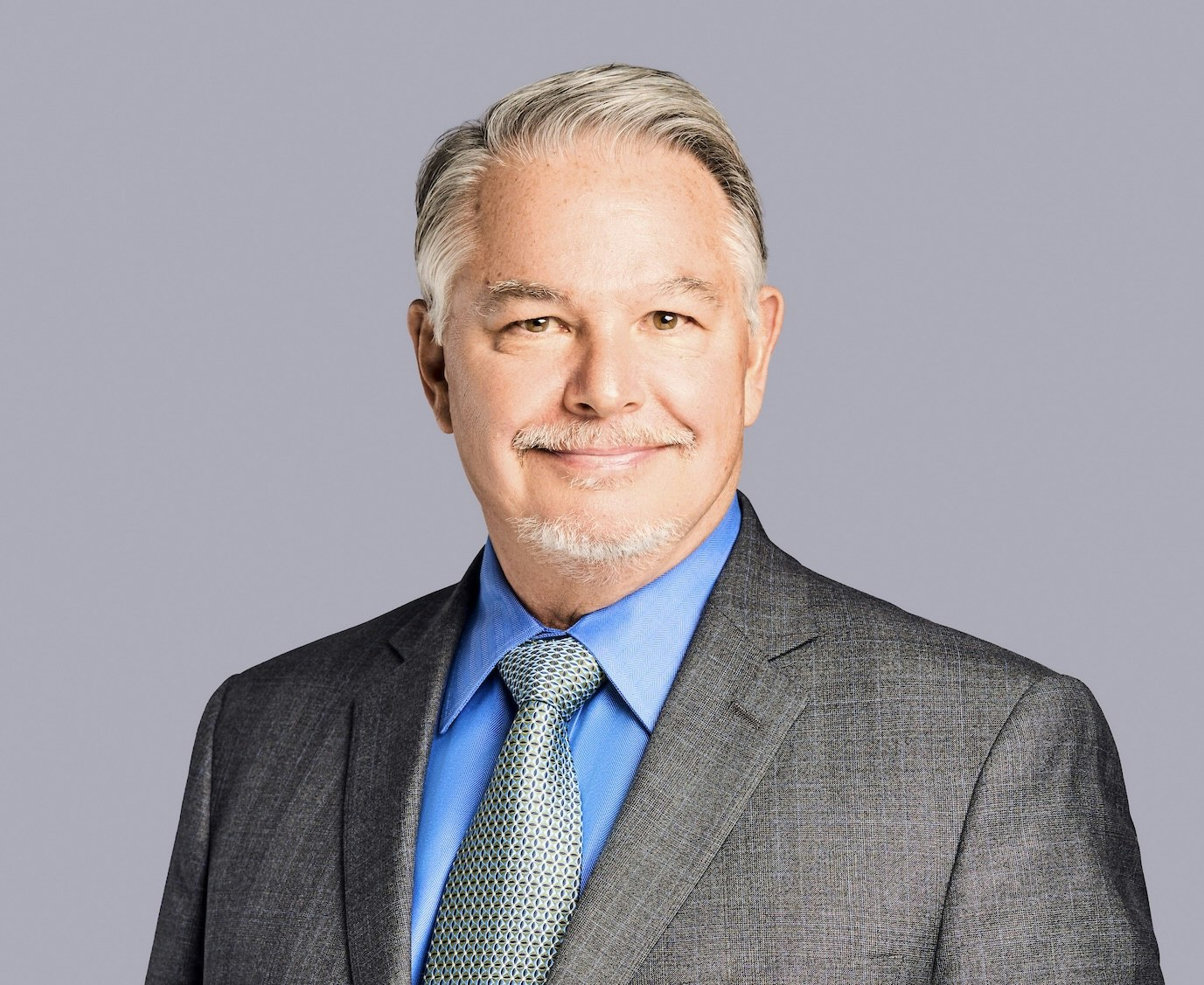 A man in a suit smiles confidently against a plain background. He has gray hair and a well-groomed beard.