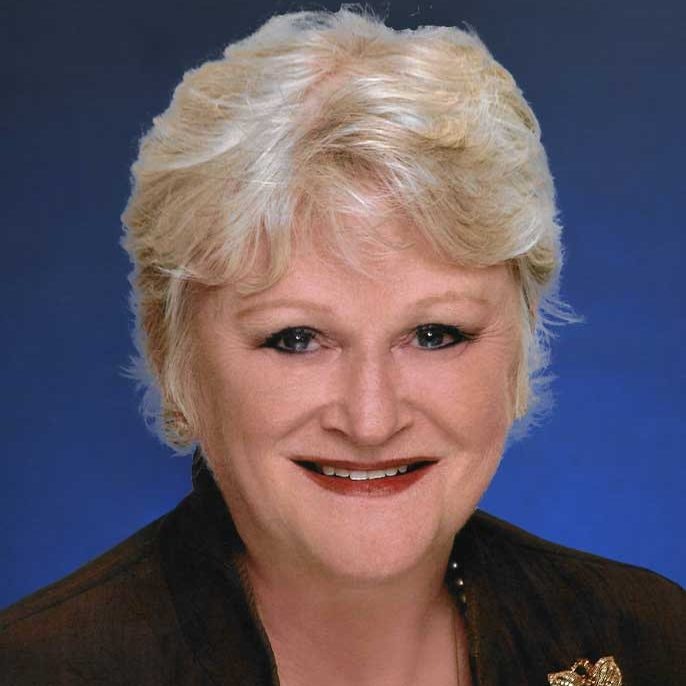A close-up portrait of a smiling person with blonde hair, make-up, and a dark outfit.