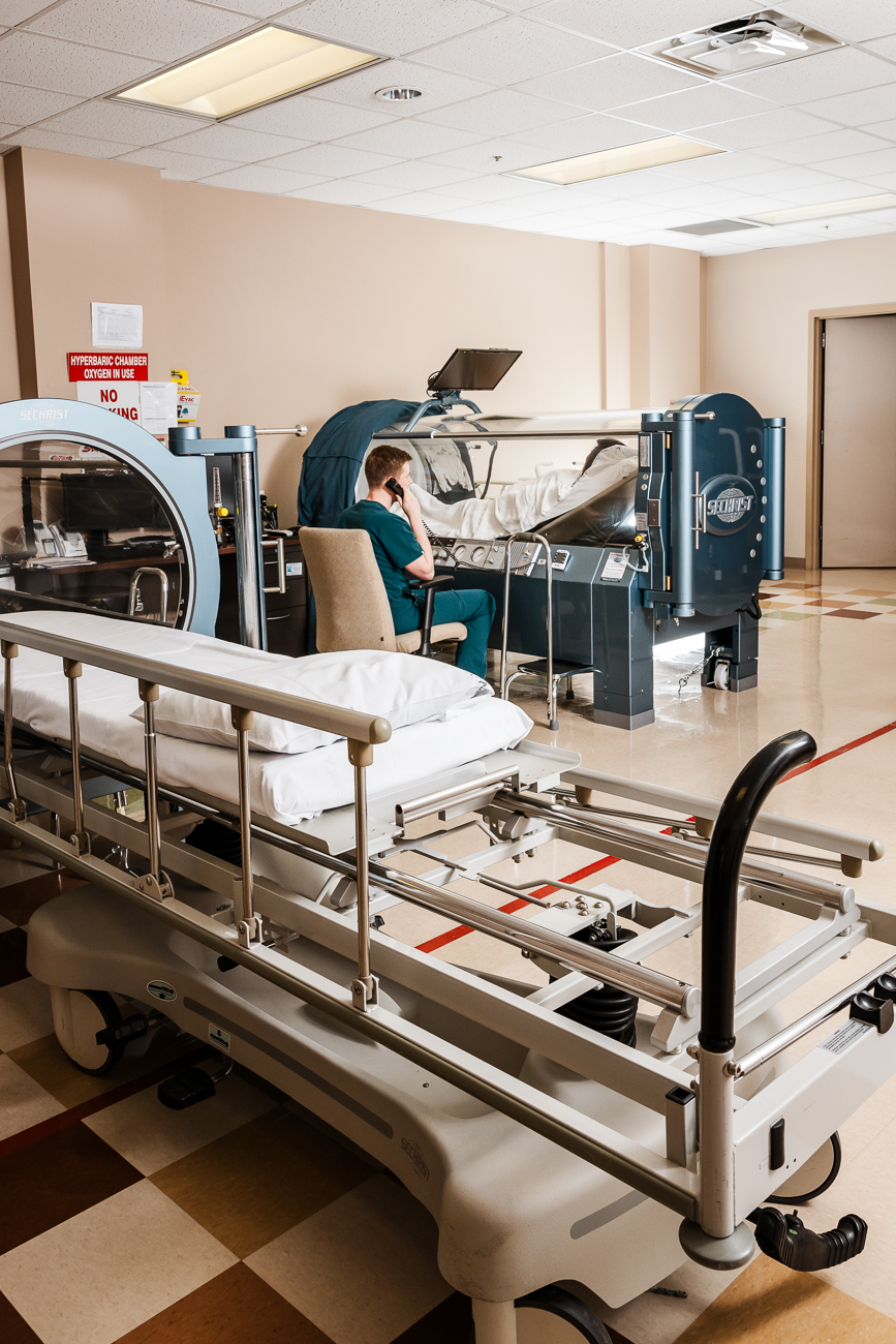 A hospital room with a hyperbaric chamber, an empty bed, and a healthcare worker on the phone.